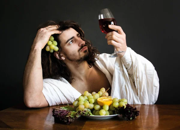 Foto medio desnudo rizado chico sentado en la mesa con frutas mira a un vaso de vino — Foto de Stock