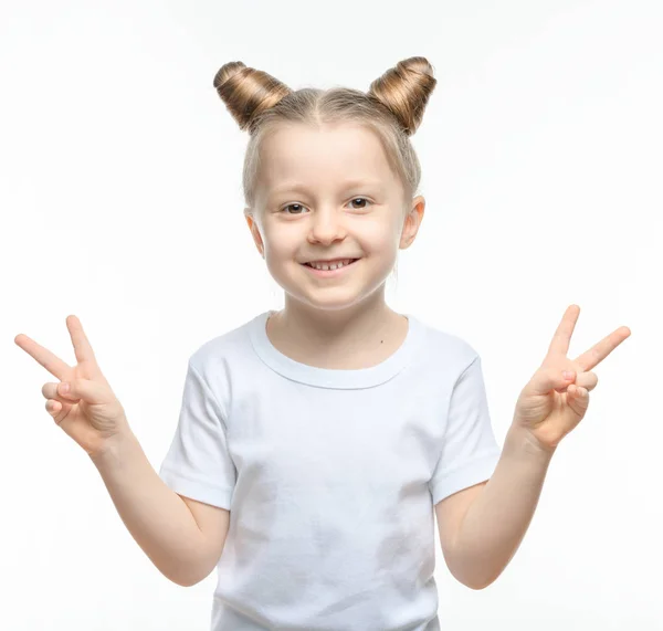 A small blonde girl in a white t-shirt smiles and shows a two-handed victory gesture on a white background. — Stock Photo, Image