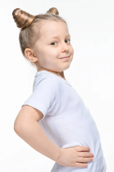 A little blonde girl in a white t-shirt with horns of hair stands sideways with her hand on her belt. — Stock Photo, Image