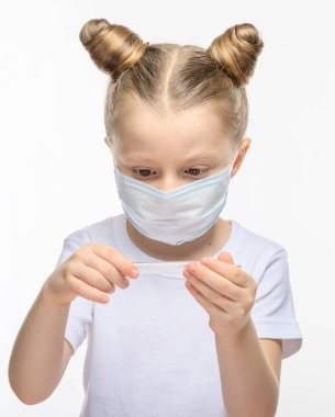 Tıbbi maskeli sarı saçlı küçük bir kız termometreye bakıyor. Tıbbi epidemiyolojik dönem, solunum hastalıkları ve yüksek sıcaklık kavramı.