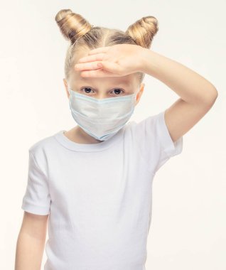 Tıbbi maskeli sarı saçlı küçük bir kız eliyle alnına dokunuyor. Tıbbi epidemiyolojik dönem, solunum hastalıkları ve yüksek sıcaklık kavramı.