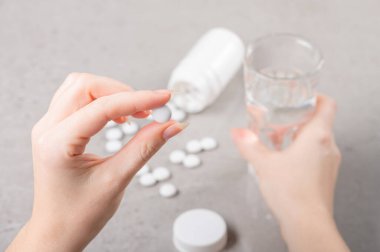Kız elinde beyaz bir tablet, vitamin ya da yiyecek takviyesi tutuyor, arka planda bir masa ve ikinci elinde bir bardak su var. Tedavi, önleme veya diyet kavramı.