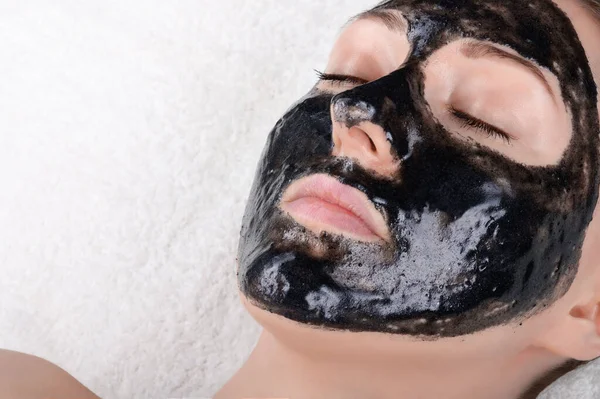 Yüzünde siyah bir maske olan güzel bir kız gözleri beyaz yumuşak bir havluya kapalı yatıyor. Aktif karbonlu siyah yüz maskesi. Güzellik salonunda ya da evde yüz bakımı, gözenekleri temizlemek..