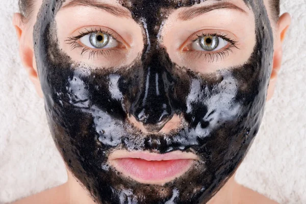 Yüzünde siyah bir maske olan güzel bir kız beyaz yumuşak bir havlunun üzerinde uzanıyor. Aktif karbonlu siyah yüz maskesi. Güzellik salonunda ya da evde yüz bakımı, gözenekleri temizlemek..