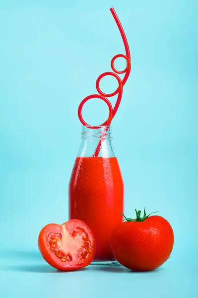 Cold tomato juice in a bottle with a beautiful straw and fresh tomatoes and slices on a blue background. The concept of an environmentally friendly and healthy drink from an organic farm.