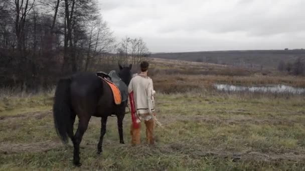 Az amerikai őslakos egy fekete lovat vezet gyeplővel a mezőn keresztül az erdő ellen.