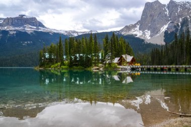 A clear reflection of tall pine trees and mountains on Emerald Lake where a wooden bridge leads to buildings on an island in the middle of the lake on a calm and cloudy day.