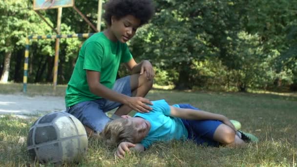Afrykański chłopiec pocieszający nastolatek przyjaciel cierpi ból leżąc na ziemi, uraz sportowy — Wideo stockowe