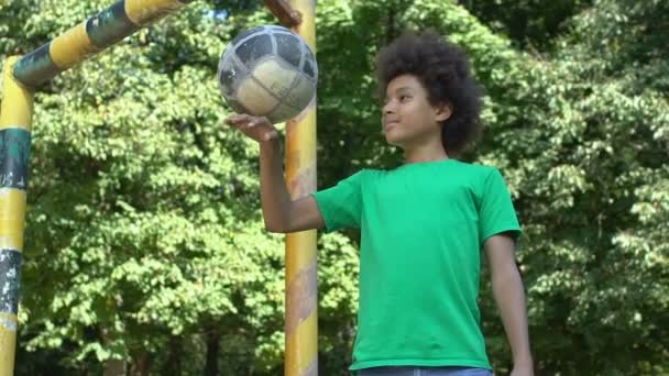 Улыбающийся чернокожий ученик играет в мяч стоя ворота стадиона, спортивная деятельность — стоковое видео