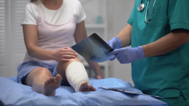Cirujano informando resultado de radiografía de hueso de rodilla a paciente femenina en envoltura de pierna elástica — Vídeos de Stock