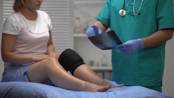 Médico explicando la radiografía del hueso de la pierna rota a la mujer en rodillera de neopreno — Vídeos de Stock