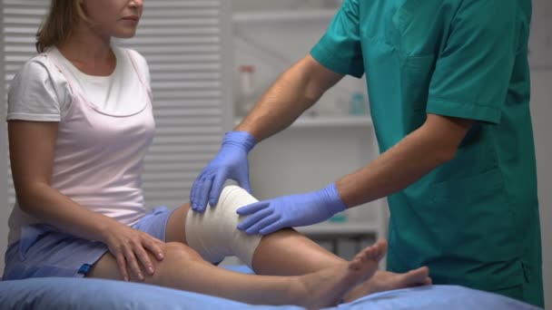 Traumatólogo revisando rodilla femenina en envoltura elástica durante el examen, trauma — Vídeos de Stock