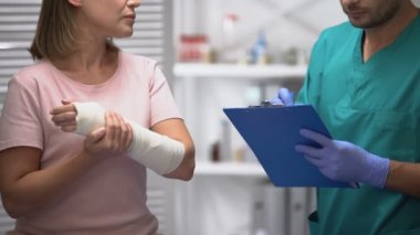 Doktor tıbbi belgeleri dolduruyor ve kadın kolunu elastik paket rehabilitasyonunda kontrol ediyor.