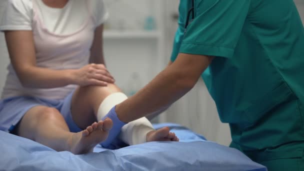 Άνδρας χειρουργός που εφαρμόζει ελαστικό περιτύλιγμα στο πόδι των γυναικών ασθενών, περίοδος αποκατάστασης — Αρχείο Βίντεο