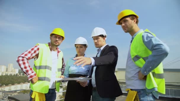Architekci omawiający plan budynku z pracownikami budowlanymi stojącymi na dachu — Wideo stockowe