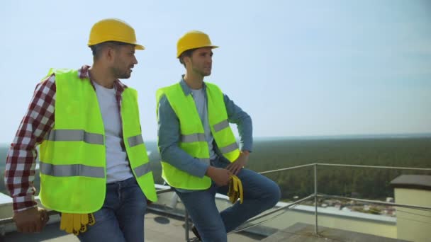 Two construction workers talking during work break on building roof, occupation — ストック動画
