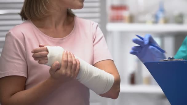 Médico consultando paciente femenina con vendaje elástico del brazo, resultado de la escritura — Vídeos de Stock
