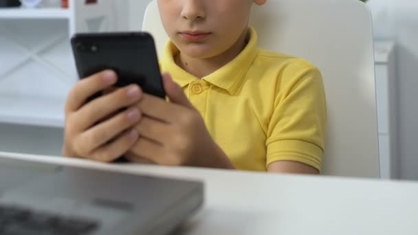 Kleiner Junge sitzt vor dem Laptop, schaut Videos auf dem Smartphone, sucht — Stockvideo