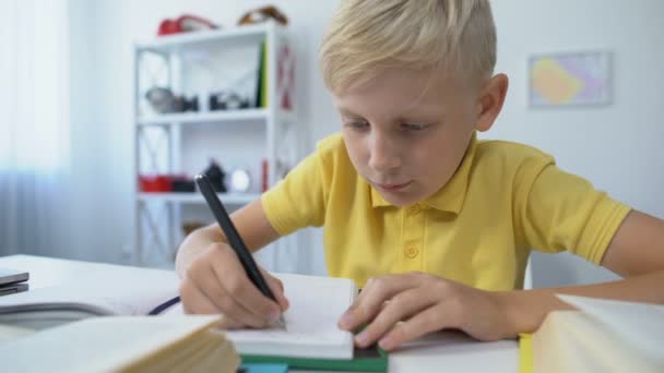 Niño varón rubio escribiendo hometask en el cuaderno y poniendo la cabeza en la mesa, aburrimiento — Vídeo de stock