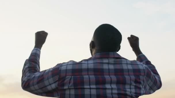 Μαύρος άνδρας σηκώνεται με τα χέρια ψηλά δείχνοντας την επιτυχία του, νικητής, μαγική ώρα, προβολή πίσω — Αρχείο Βίντεο