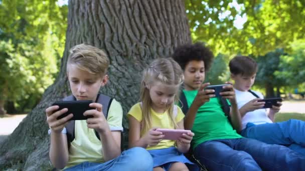 Kinder spielen Geräte, sitzen unter einem Baum im Park, Junge lächelt in die Kamera, App — Stockvideo