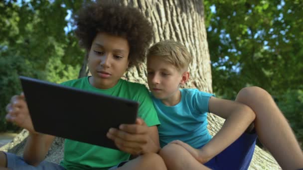 Мальчики играют на планшете, сидят под большим деревом в парке, друзья проводят свободное время — стоковое видео