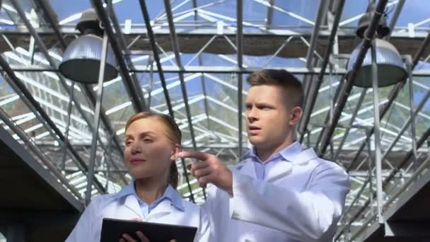 Man and lady in white suits walking in greenhouse, analyzing experiment results — Stock Video