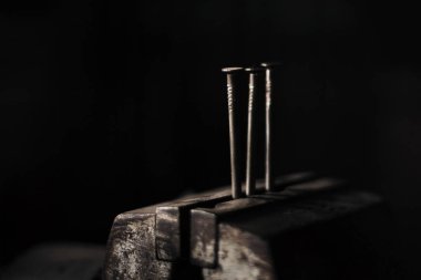 Image - 3 silver iron nails standing in bench vise iluminated with natural light on black and dark background. Heads of nails in darkness.