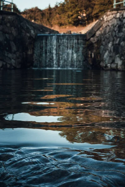 Foto vertical de pequeno fluxo de cachoeira que flui da lagoa (lago) com o pôr do sol no fundo. Cachoeira com água azul cristalina e parede de pedra com ondas - reservatório no outono (outono ). — Fotografia de Stock