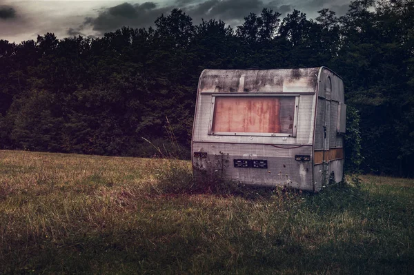 Abandoned and rusty small travel trailer in the meadow with forest on background and dark cloudy sky. Toned photo with violet colors of old and damaged caravan after season.