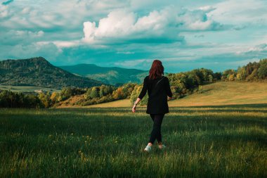 Kıvırcık saçlı mutlu ve genç, kızıl saçlı kız (manken) arka planda bahar ve ormanla birlikte çayırda yürüdü. Doğadaki kadın - hayattan zevk alıyor.