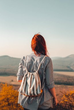 Genç gezgin ve maceraperest kızıl saçlı kız arka planda tepeler olan vadide mükemmel manzaranın tadını çıkarıyor. Gün batımı çekimi. Sırt çantalı bir kadın vahşi doğayı keşfediyor. Arkadan çekilmiş bir fotoğraf..