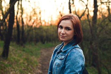Gün batımında mavi kot ceketli, kahverengi gözlü, genç ve güzel bir kızıl kadın portresi. Yatay çekim - Yumuşak ciltli kız (manken) ve orman yolunda kameraya poz veren güzel gülümseme.