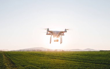 Beyaz dron gün batımında buğdayla yeşil arazide uçuyor. Drone Quad helikopteri, arka planda tepelerle çayırların üzerinden güneş ışınlarıyla uçuyor..