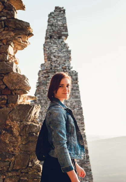 Genç ve güzel esmer kız (kadın) portresi, günbatımında antik harabelerde güzel bir gülümseme ile. Büyük siyah gözlü, mavi kot ceketli ve kameraya poz veren bir modelin dikey fotoğrafı...
