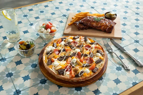 Pan πίτσα με κρέας, πατάτα, λαχανικά toppings και χοιρινό παϊδάκι και πλευρικό πιάτο σε ένα τραπέζι Εικόνα Αρχείου