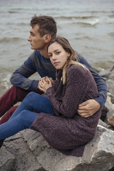 Happy thoughtful couple sitting on a rock beach near sea hugging — Stock Photo, Image