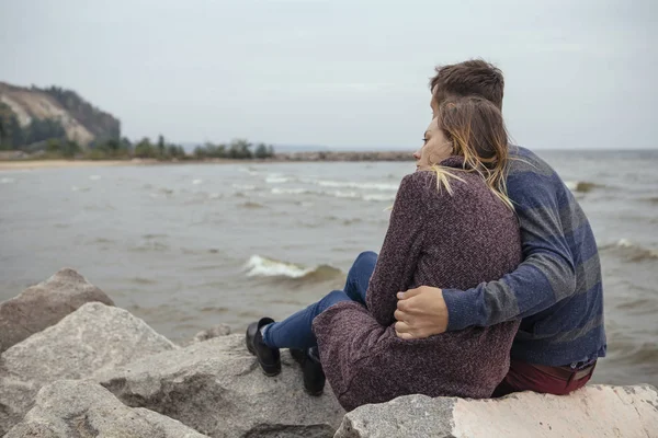 Feliz pareja pensativa sentada en una playa de rocas cerca de abrazos al mar —  Fotos de Stock