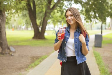 Sevimli güzel ince beyaz esmer kadın Parkı'nda yaz gün içme kırmızı limonata bir yolda rahat kıyafet (etek, tişört ve kot ceket). O tam bir sevinç, gülümseyen ve eğlenin