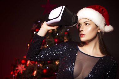 Noel şapka ve seksi Abiye Vr gözlükle sevimli gülümseyen kadın. Bir arka plan üzerinde kırmızı ışık ile yeni yıl ağacı