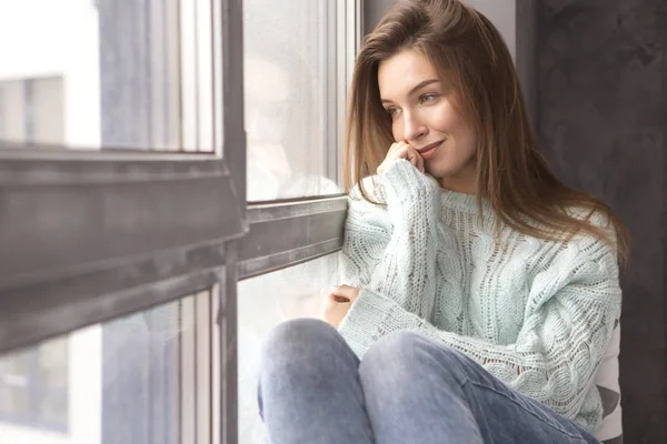 Симпатичная взрослая молодая женщина, сидящая у окна в повседневной одежде — стоковое фото