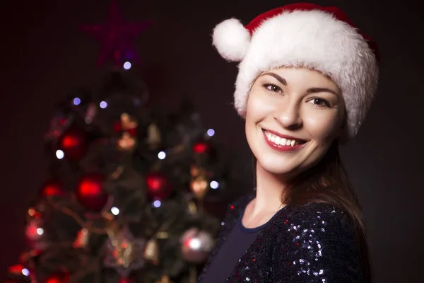 逗人喜爱的微笑的妇女在圣诞节帽子和晚上性感的礼服站立在新年树附近以红灯在背景 — 图库照片