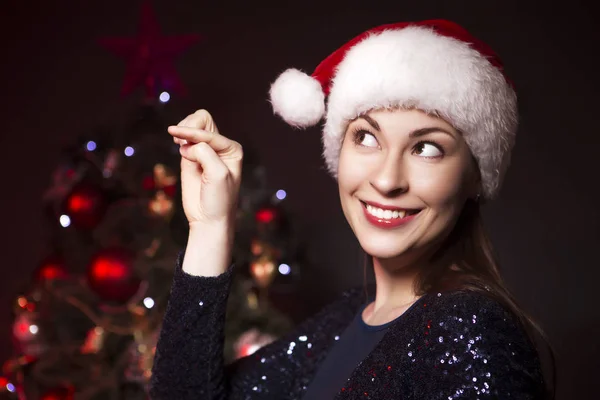 逗人喜爱的微笑的妇女在圣诞节帽子和晚上性感的礼服站立在新年树附近以红灯在背景 — 图库照片