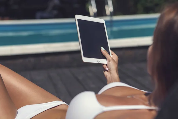 Sexy brunette caucasian woman get tan near swimming pool in a white bikini. She young and fit with healthy skin. Girl reading or working on a tablet, focus on it