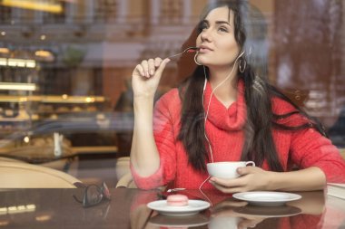 Şirin esmer bir mesafe (serbest) çalışma, kafede oturup, kahve içme ve yemek macaron boyutu kadın artı. O kırmızı kazak giyen dinlemek müzik ve ona telefon iletileri yazma