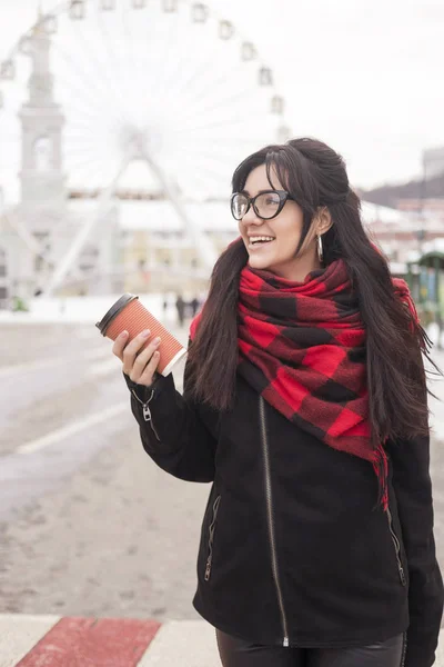 可爱的年轻白种黑发女学生在欧洲城市街道散步 她穿着拉丁大衣和格子红围巾 寒冷的冬天天气 妇女喝咖啡从一个到去的杯子 — 图库照片