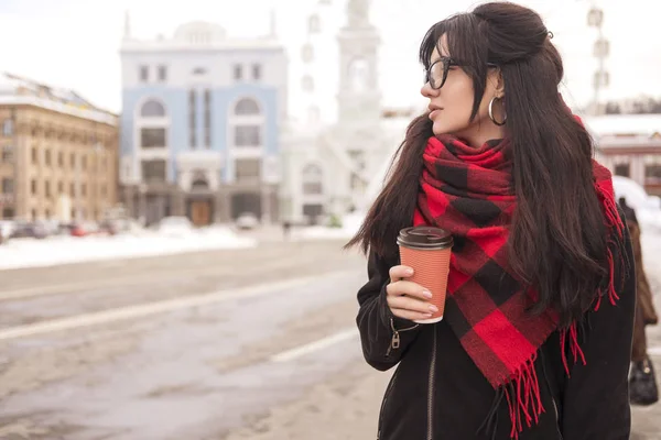 可爱的年轻白种黑发女学生在欧洲城市街道散步 她穿着拉丁大衣和格子红围巾 寒冷的冬天天气 妇女喝咖啡从一个到去的杯子 — 图库照片