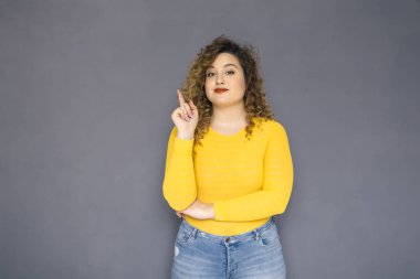 Şirin esmer artı boyutu kadın sarı kazak ve bir nötr gri arka plan üzerinde ayakta kot kıvırcık saçlı. O, yüzünde düşünceli duygu düşünüyordum. Kopya alanı