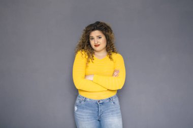 Şirin esmer artı boyutu kadın sarı kazak ve bir nötr gri arka plan üzerinde ayakta kot kıvırcık saçlı. O bir şey yüzünde mutsuz duygular şüphe