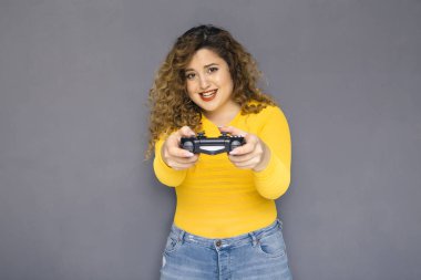 Şirin esmer artı boyutu kadın sarı kazak ve bir nötr gri arka plan üzerinde ayakta kot kıvırcık saçlı. O bir joystick konsol oyunları oynarken, tutar